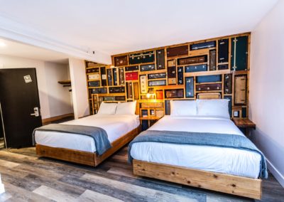 View of a room with two beds, wood looking floors and a back wall that looks like a bunch of suitcases.