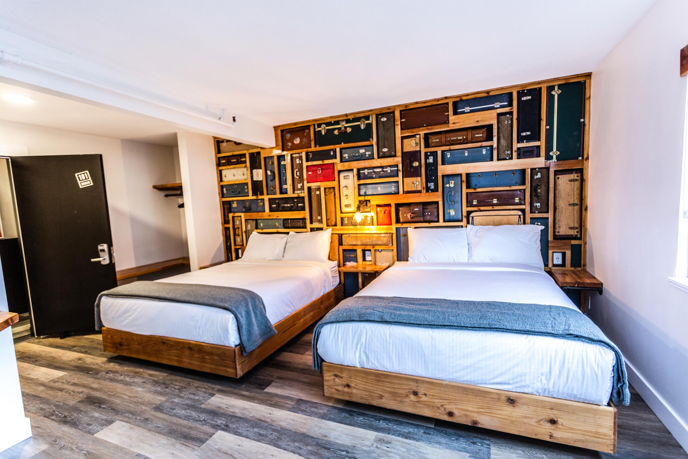 View of a room with two beds, wood looking floors and a back wall that looks like a bunch of suitcases.